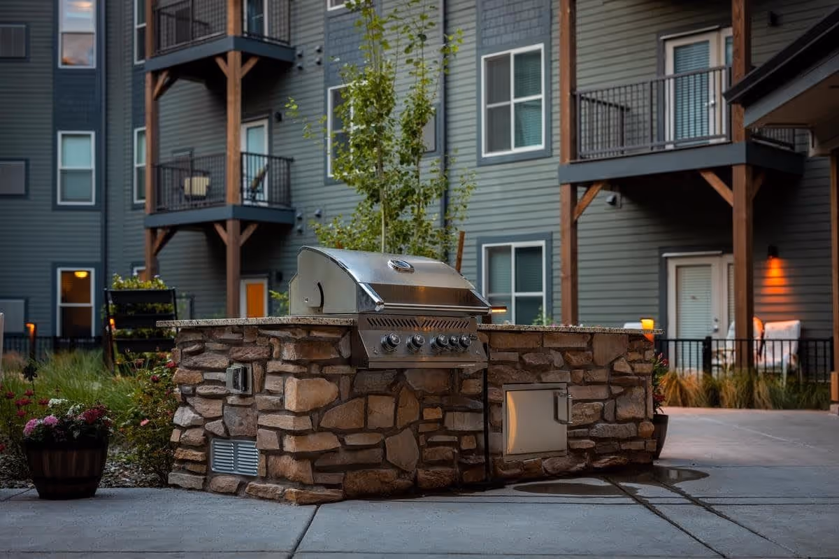 Outdoor stone grill station with a stainless steel barbecue grill in the courtyard of a senior living facility. The background shows a multi-story building with balconies, windows, and patio doors, along with some plants and outdoor seating.