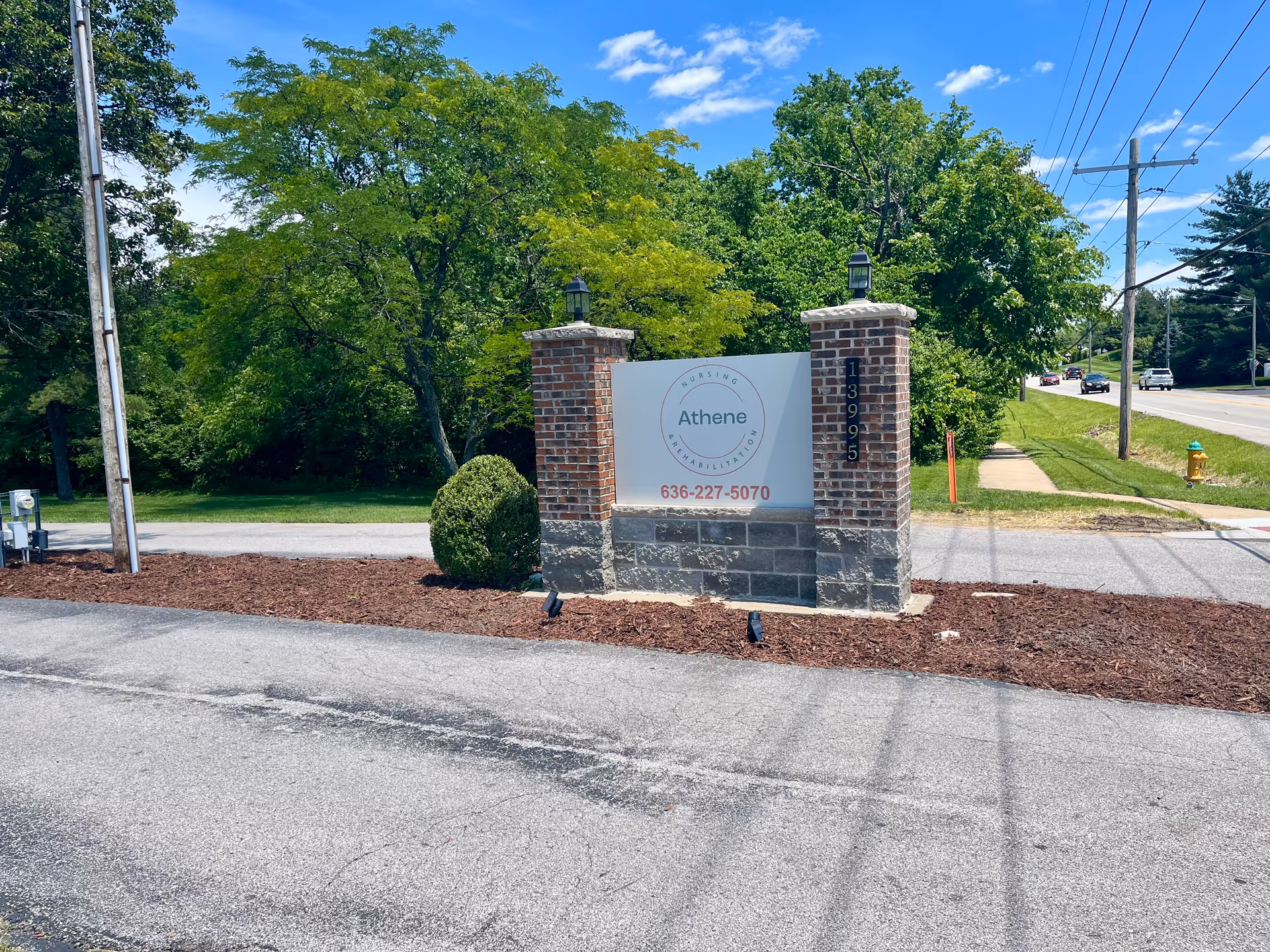 Outdoor view of a brick and stone sign for Athene Nursing and Rehabilitation with the phone number 636-227-5070, surrounded by greenery and trees, next to a road with cars and utility poles under a blue sky with some clouds.