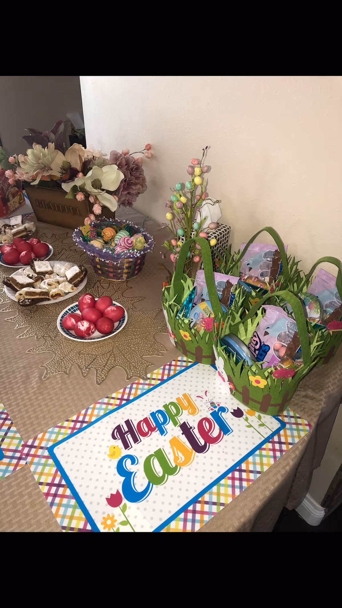 A decorated table with Easter-themed items including two green baskets filled with candy, plates of red dyed eggs and pastries, a floral centerpiece, and a colorful placemat that says 'Happy Easter'.