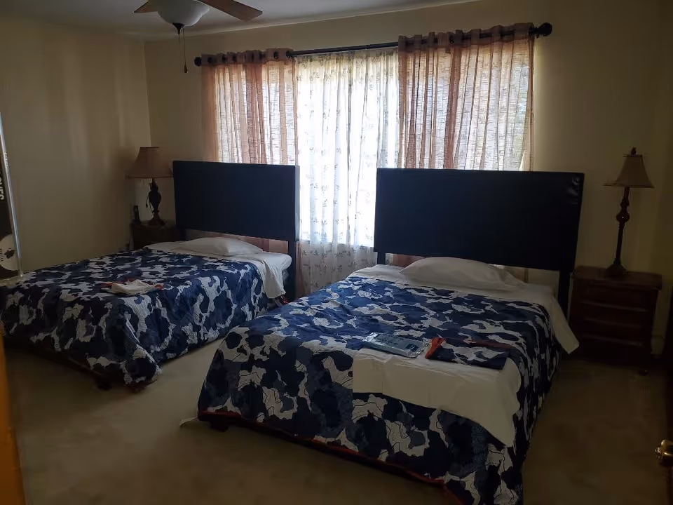 A bedroom with two twin beds featuring dark headboards and blue floral patterned bedspreads. Each bed has a pillow and a folded towel or cloth on top. There are two bedside tables, each with a lamp, and a window with sheer and brown curtains letting in natural light.