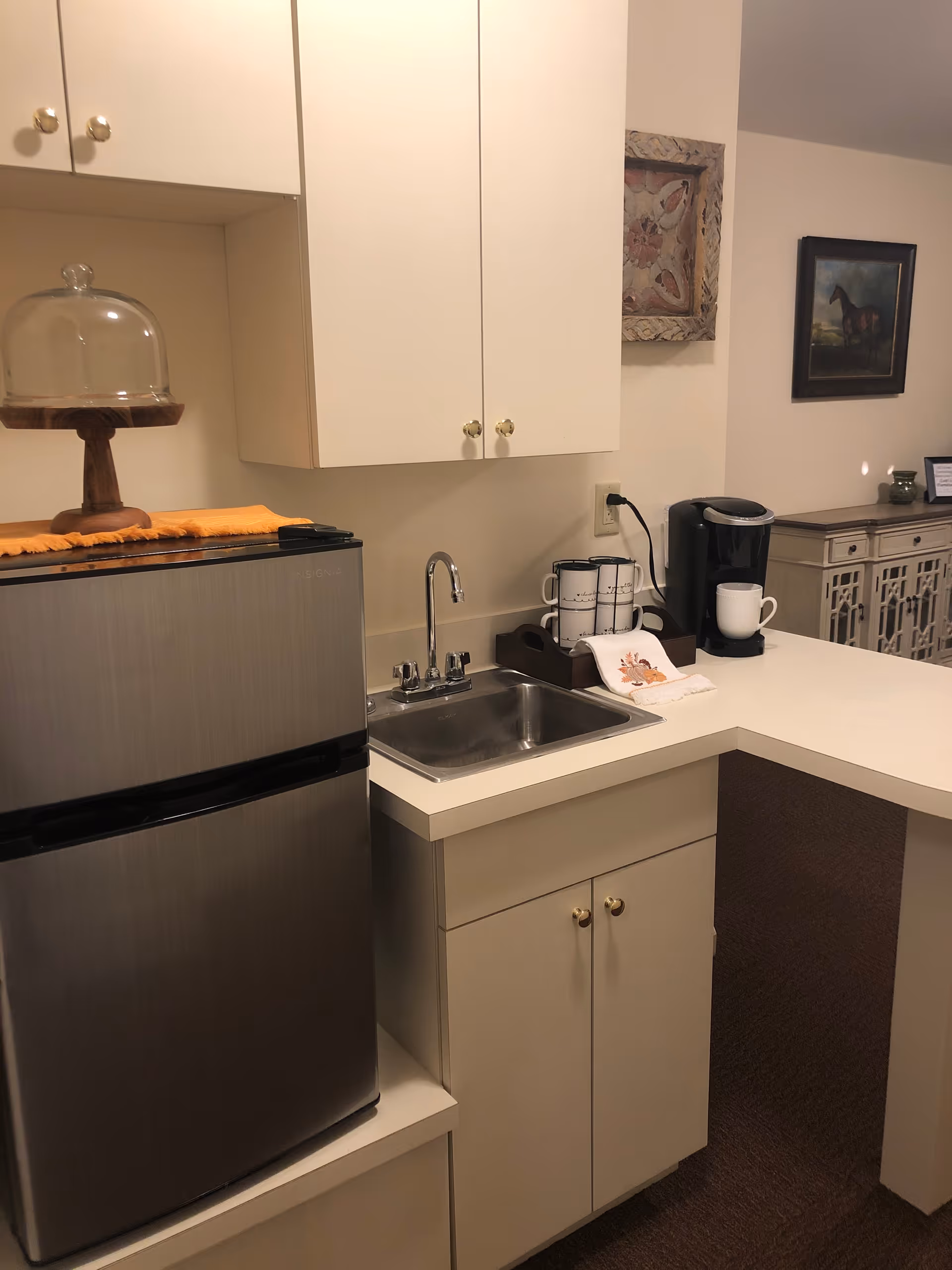 Small kitchenette with a stainless steel mini fridge, sink, coffee maker, cabinets, and countertop decor.