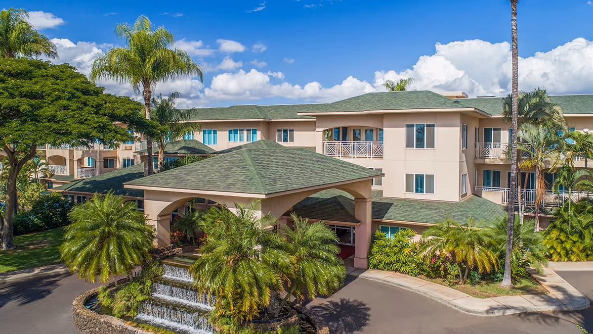 Exterior view of Holiday Kalama Heights facility showing a multi-story building with green roofs, surrounded by palm trees and lush landscaping under a partly cloudy blue sky. There is a covered entrance with a tiered water fountain and tropical plants in front.