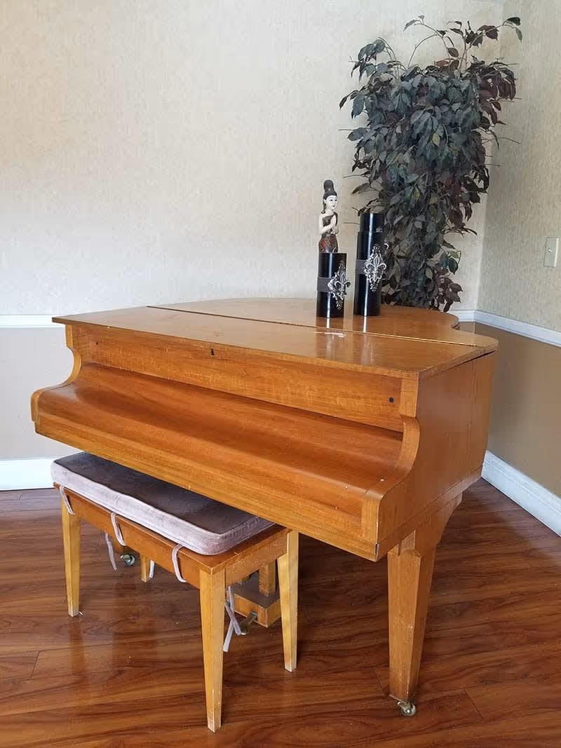 A wooden baby grand piano with a matching wooden bench that has a cushioned seat, placed on a polished wooden floor. On top of the piano are two black decorative candle holders with silver embellishments and a small figurine. In the background, there is a tall artificial plant against a beige wall with white trim.