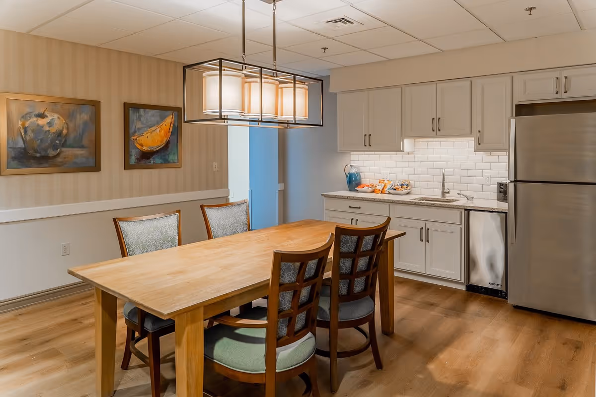 A dining area with a wooden table and four chairs with patterned cushions. Above the table hangs a modern rectangular light fixture with four lamps. On the right side, there is a kitchen area with white cabinets, a stainless steel refrigerator, a small sink, and a countertop with snacks and a blue pitcher. The walls have light-colored wallpaper with two framed paintings of fruit.