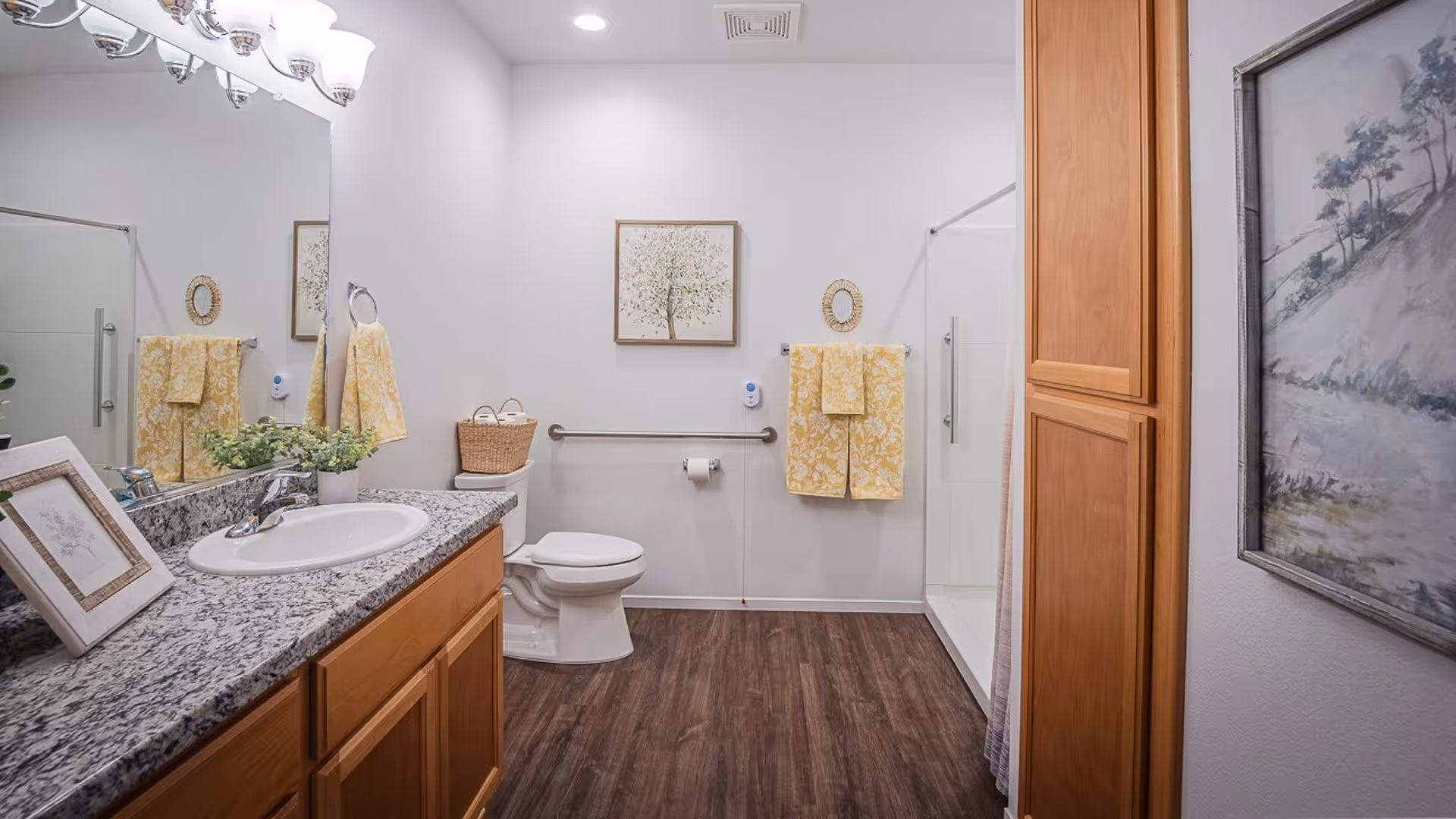 A clean and well-lit bathroom with a granite countertop sink, wooden cabinets, a toilet with a basket on top, a walk-in shower with a glass door, yellow patterned towels hanging on the wall, and framed artwork on the walls.