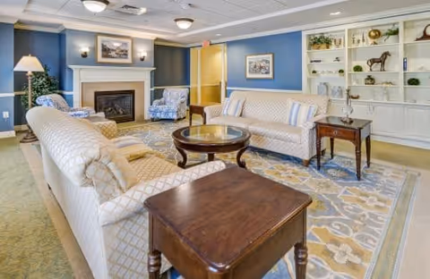 A cozy living room with two patterned sofas, two armchairs, a round wooden coffee table, and side tables. The room features a fireplace with a framed picture above it, blue walls, built-in white shelves with decorative items, and a large patterned area rug covering the floor.