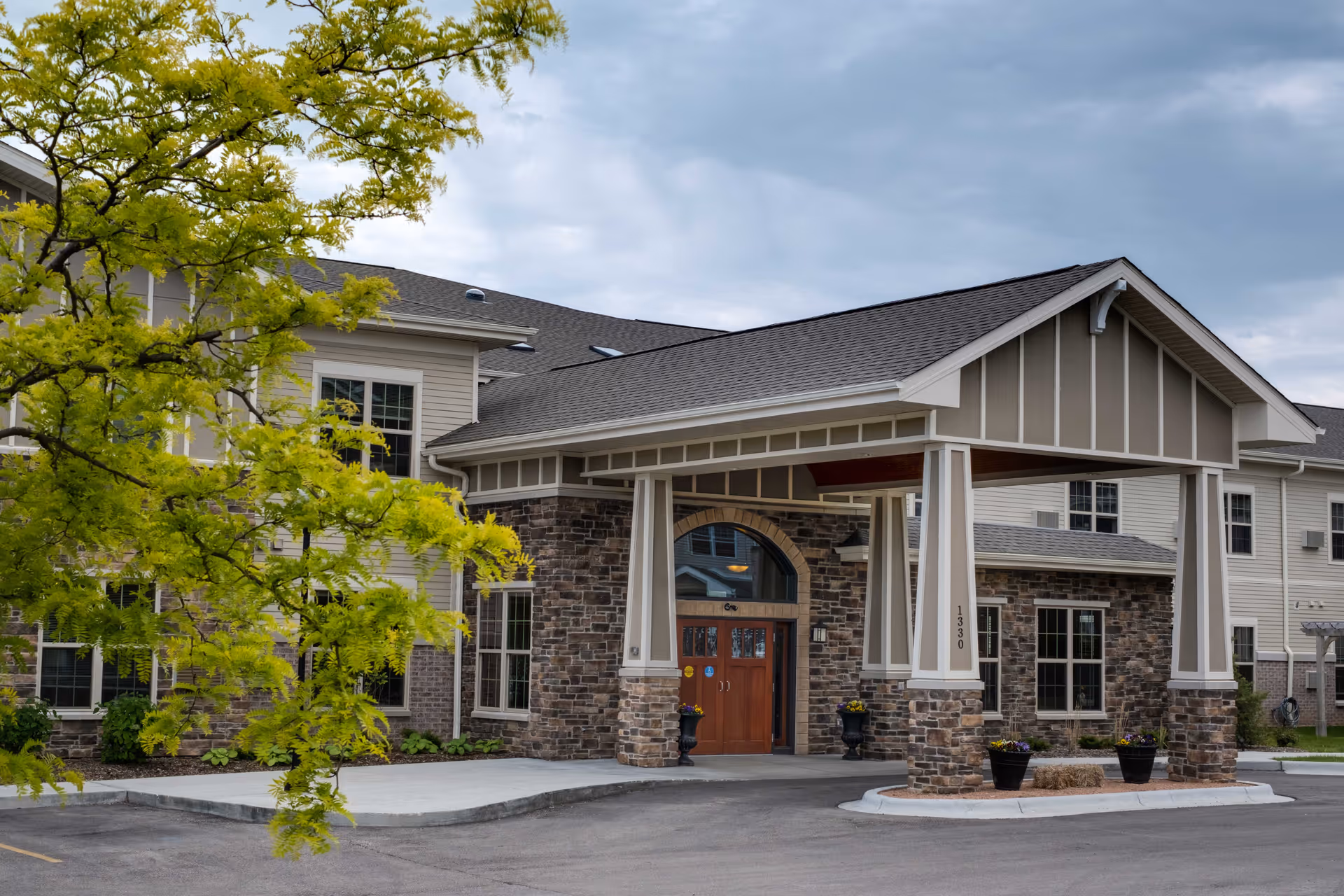 Exterior view of Lincoln Village Senior Campus showing the main entrance with a covered driveway supported by stone and white pillars. The building has beige siding with stone accents and several windows. A tree with green leaves is visible on the left side under a cloudy sky.