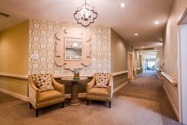 A cozy seating area in a hallway with two beige armchairs featuring patterned cushions, a round wooden table with a decorative item on top, a large ornate mirror on the wall, and a patterned wallpaper behind the seating. The hallway extends into the distance with doors and handrails along the walls.