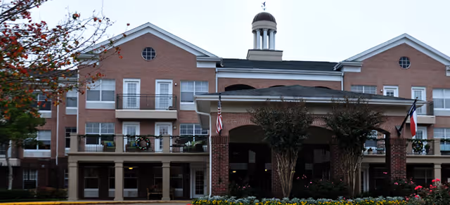 Front exterior view of a large brick senior living facility building with multiple windows, balconies decorated with wreaths, a covered entrance supported by brick columns, and two flags displayed near the entrance. Trees and flower beds are visible in the foreground.