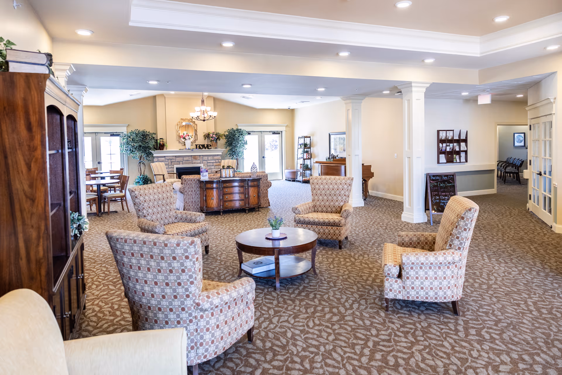 Bright common lounge in a senior living facility with patterned armchairs around a central coffee table, a fireplace and dining area in the background.