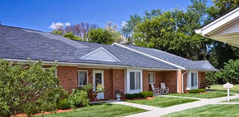 Single-story brick senior living building with front porches, chairs, a sidewalk and manicured lawn under a blue sky.