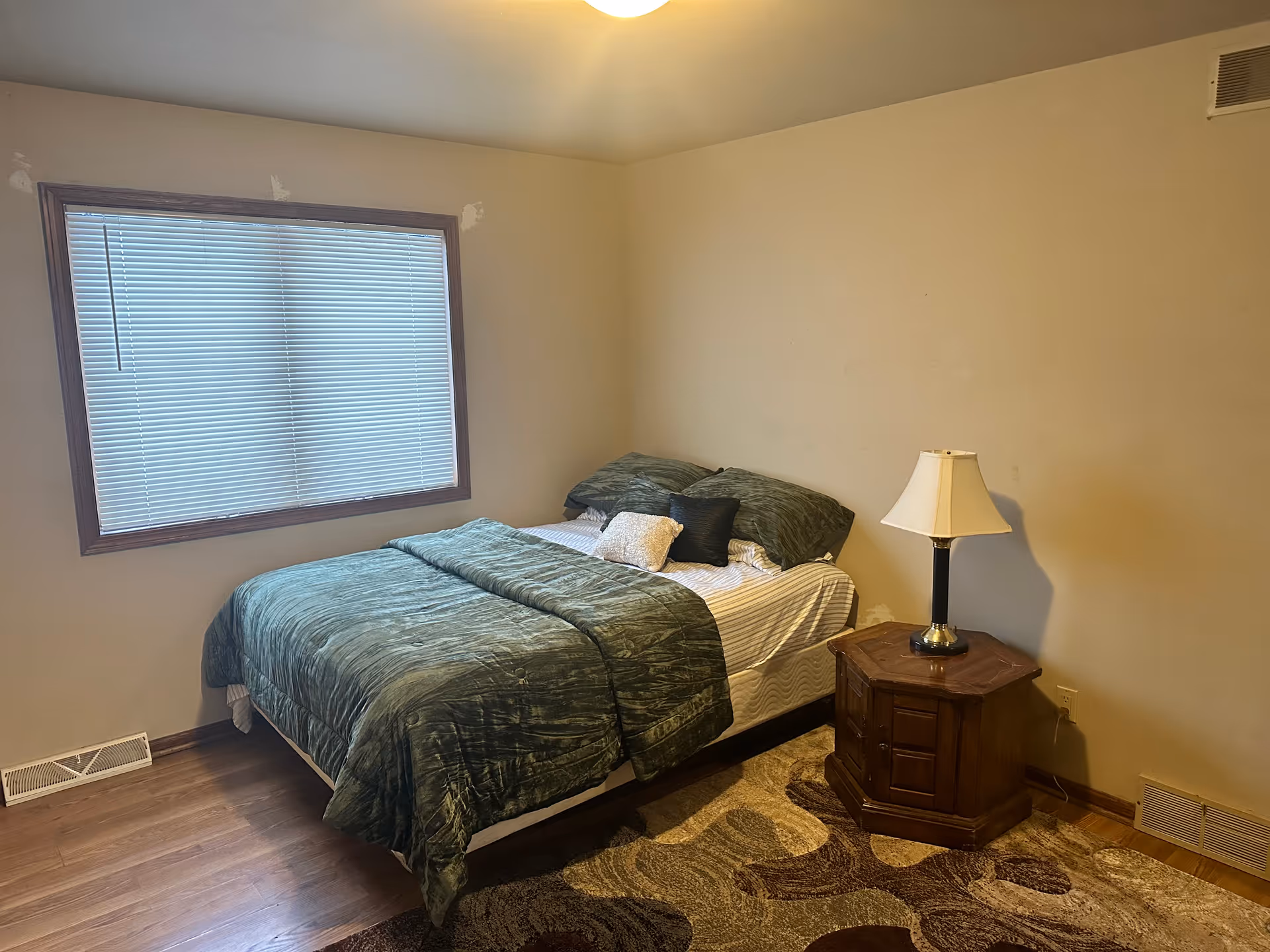 A bedroom with a bed covered in green bedding and multiple pillows. There is a wooden nightstand with a lamp on it next to the bed. The room has beige walls, a window with closed blinds, and a patterned area rug on the wooden floor.