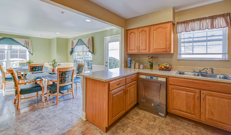 A bright kitchen area with wooden cabinets, a stainless steel dishwasher, and a double sink under a window with blinds. Adjacent to the kitchen is a dining area with a table covered by a blue tablecloth and surrounded by wooden chairs with floral upholstery. The dining area has large windows with valances and a door leading outside.