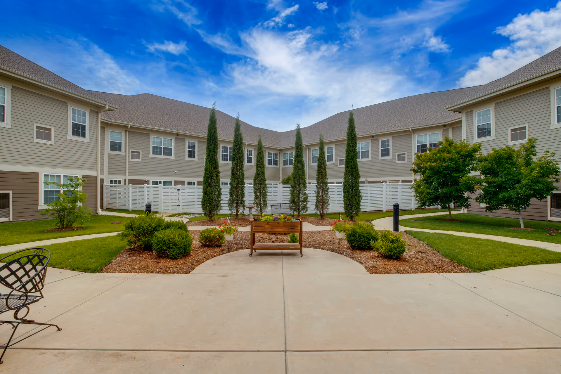Courtyard with landscaped planting beds, tall columnar trees and a bench in front of a two-story beige senior living building under a blue sky.