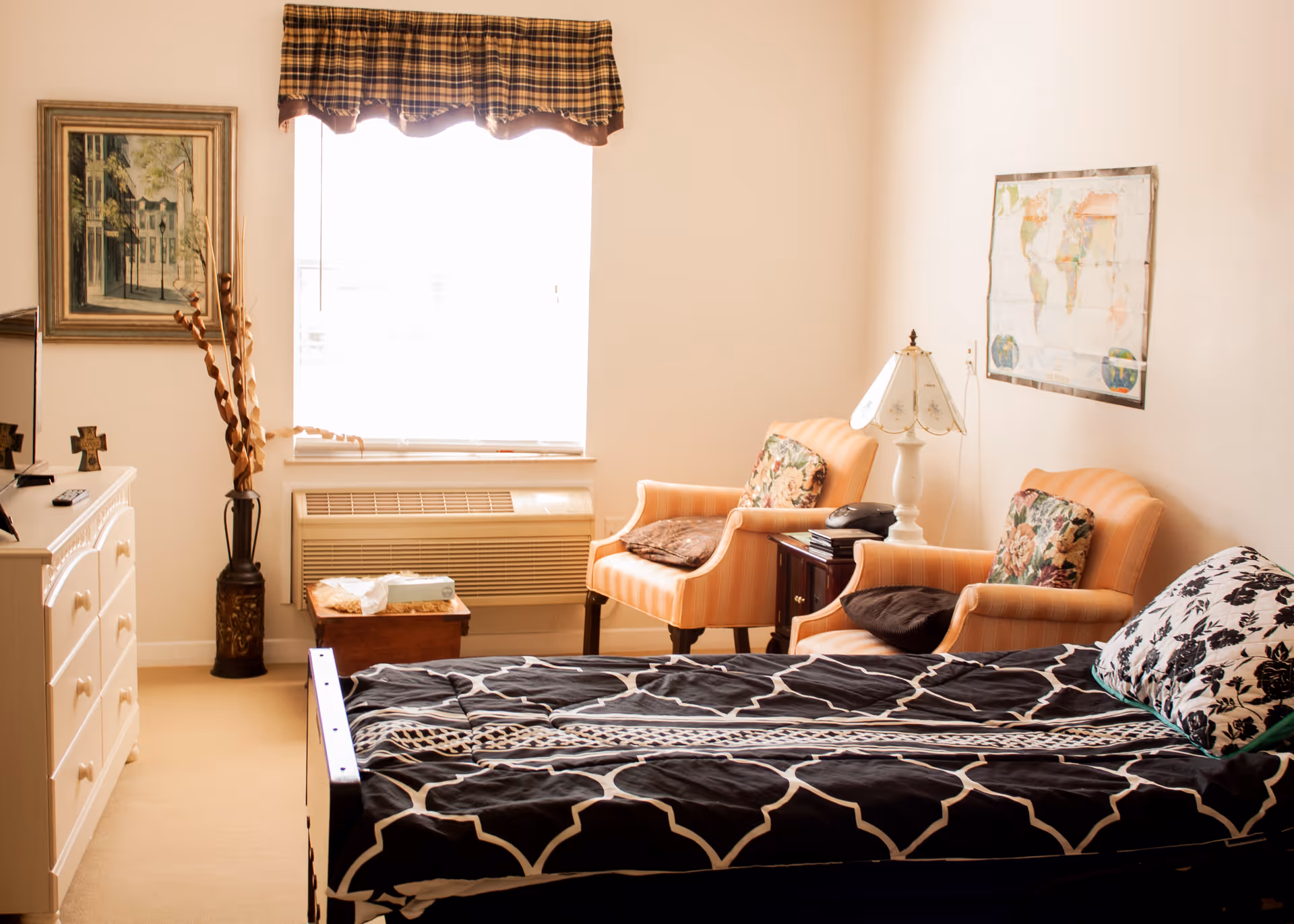A cozy bedroom in an assisted living facility featuring a single bed with black and white patterned bedding, two orange armchairs with floral cushions, a small wooden side table with a lamp and telephone, a window with a plaid valance, a dresser with a TV and decorative items, a framed painting on the wall, and a world map poster.