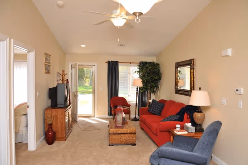 A cozy living room with beige walls and carpet. The room features a red sofa with dark cushions, a blue armchair, a wooden side table with a lamp, and a wicker coffee table. There is a large mirror on the wall above the sofa and a tall potted plant next to a floor lamp near the window with dark curtains. A wooden TV stand with an older-style television is positioned against the left wall, near an open door leading to a bedroom. The back door is open, showing a green outdoor area.