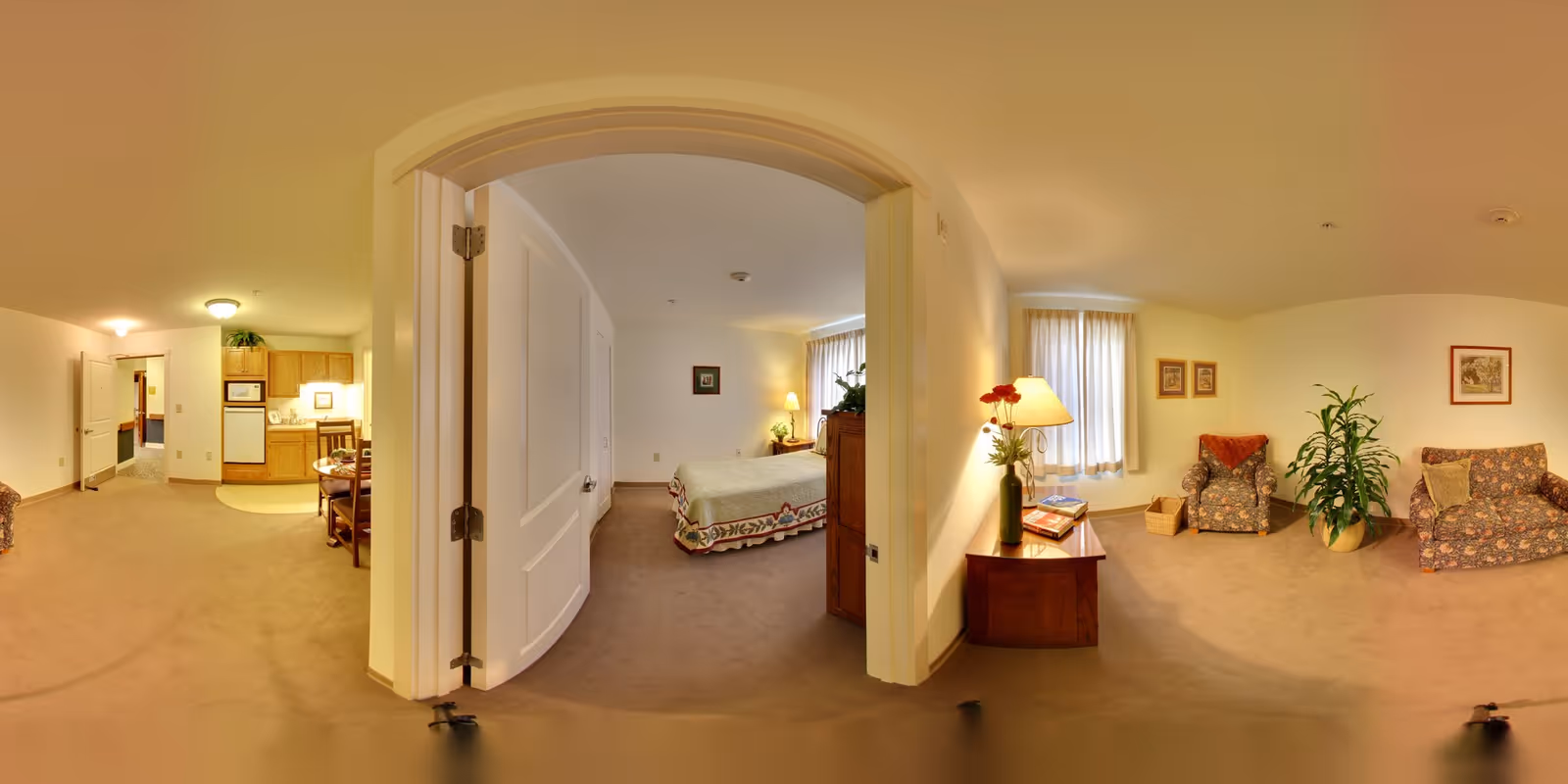 A panoramic view of an assisted living apartment showing a small kitchen area with wooden cabinets, a dining table with chairs, a bedroom with a bed and dresser, and a living room area with two floral armchairs, a floral sofa, a side table with a lamp and flowers, and framed pictures on the walls. The space is carpeted and well-lit with natural and artificial light.