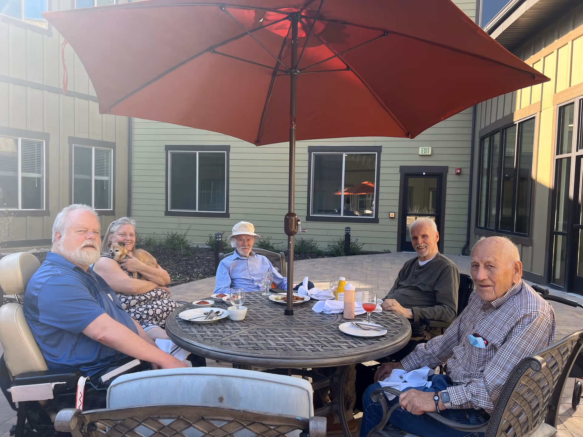 Five elderly people sitting around a round outdoor table with a large red umbrella, enjoying a meal together on a patio area outside a senior living facility. One woman is holding a small dog. The building exterior with windows and doors is visible in the background.