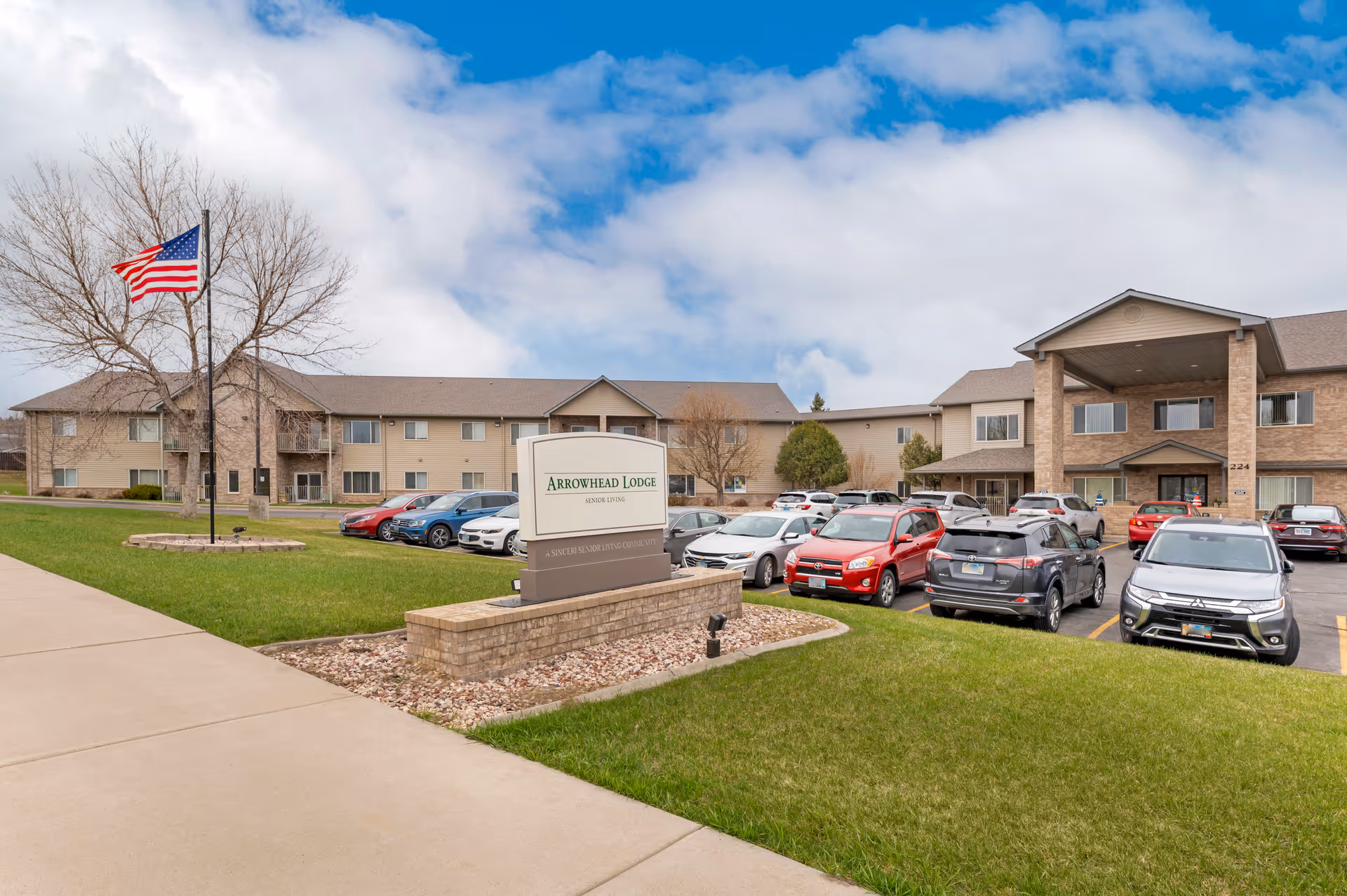 Exterior view of Arrowhead Lodge Senior Living facility showing a two-story building with a covered entrance, several parked cars in the parking lot, a green lawn, a sidewalk, and an American flag on a flagpole.