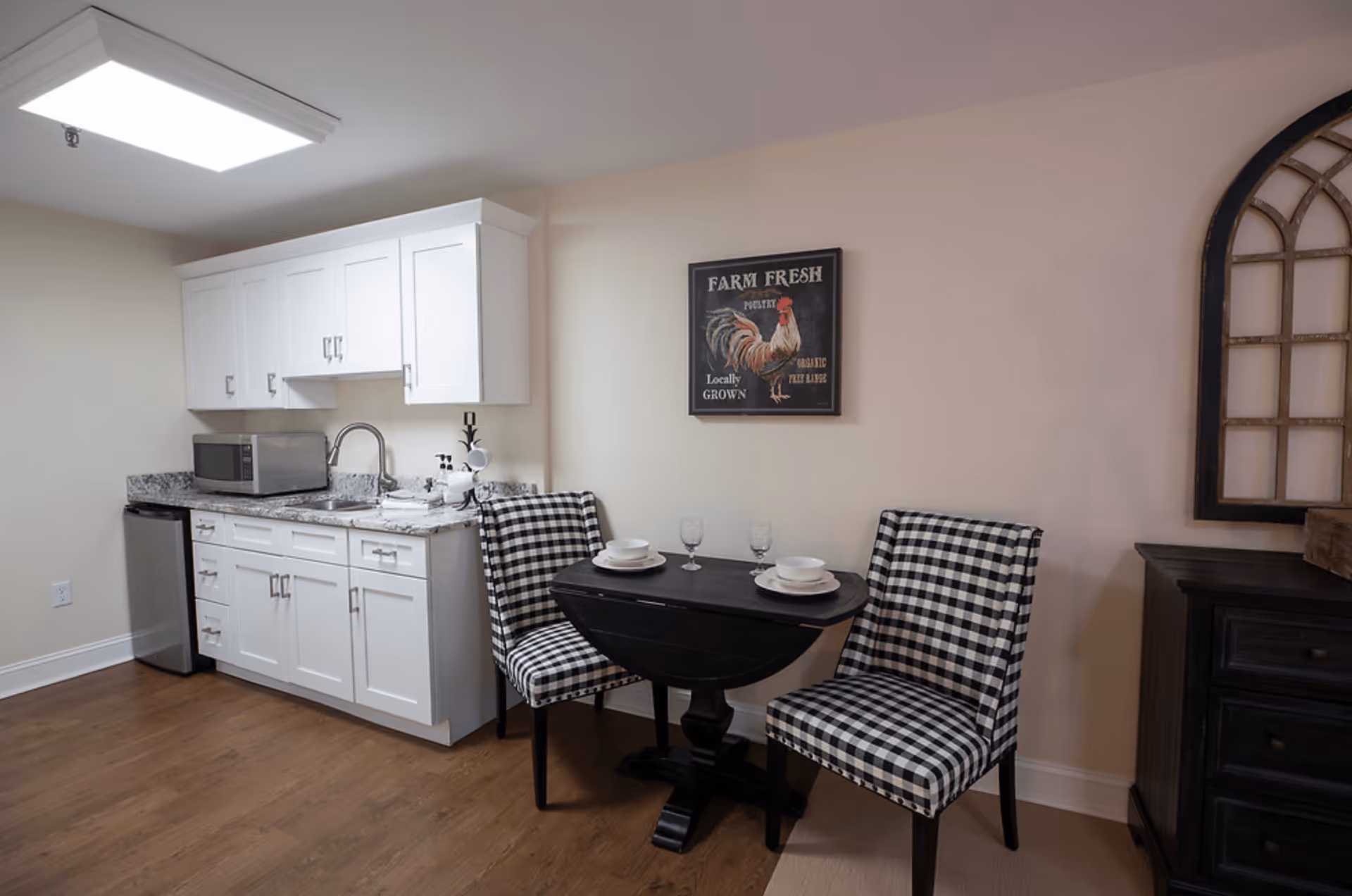A small kitchen area with white cabinets, a granite countertop, a microwave, and a mini fridge. Next to the kitchen is a small black dining table set for two with black and white checkered chairs. A framed picture of a rooster with the text 'Farm Fresh' hangs on the wall above the table. The floor is wooden, and there is a decorative window frame on the right side.