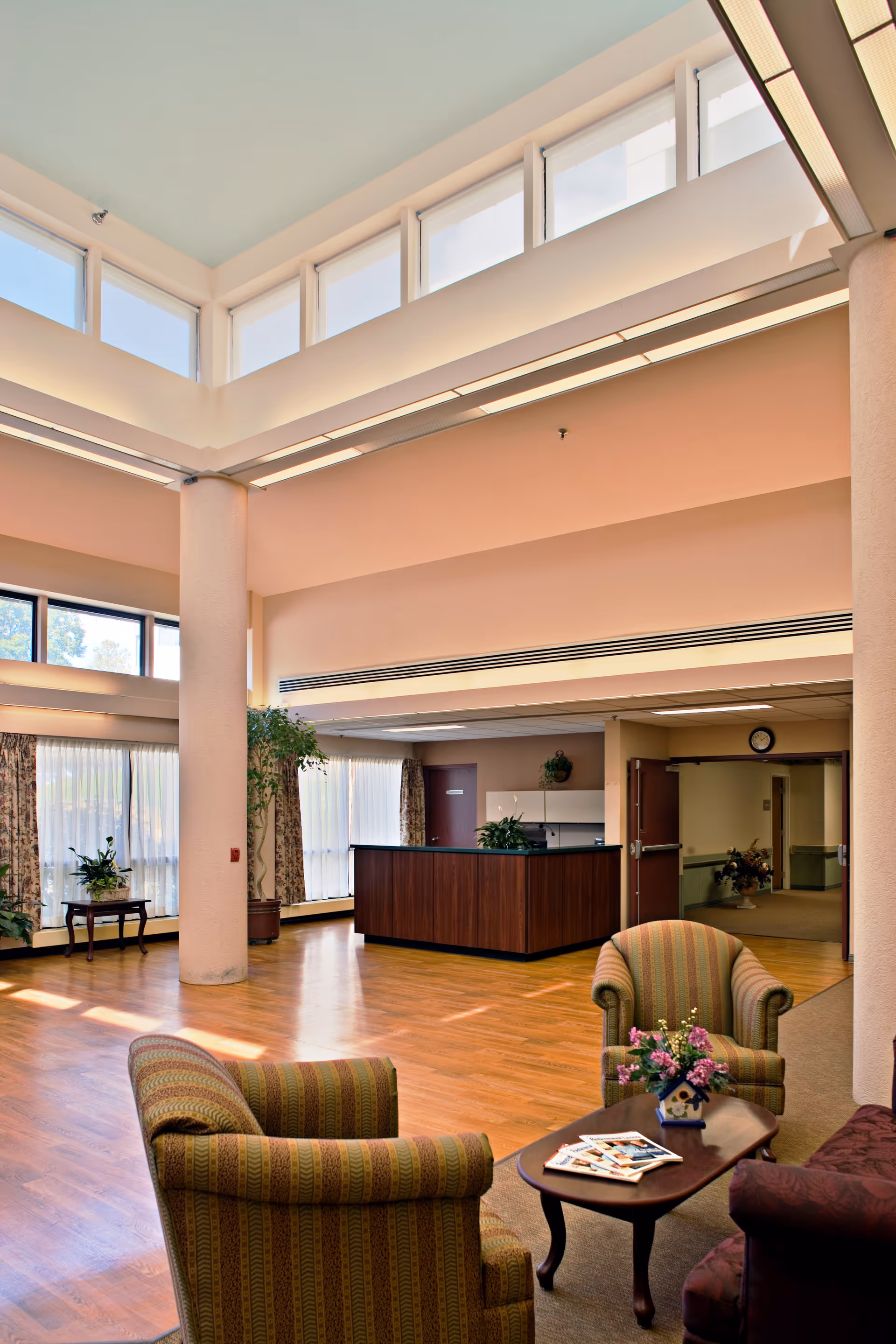 A spacious and well-lit common area with high ceilings and large windows. The room features two upholstered armchairs and a small wooden coffee table with a flower arrangement and magazines. In the background, there is a reception desk with plants and a hallway leading to other rooms.