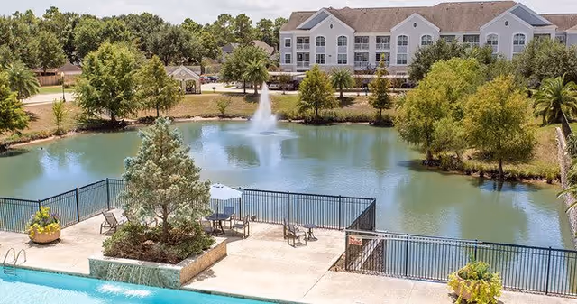View of a serene outdoor area at The Grand Parkway featuring a swimming pool with lounge chairs and umbrellas, a fenced patio area, a pond with a water fountain, and a large multi-story building in the background surrounded by trees and greenery.