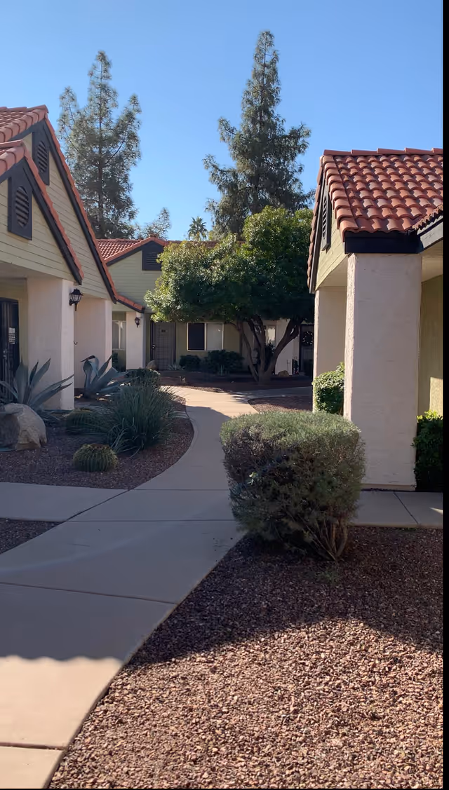 Paved courtyard walkway between stucco single-story buildings with red tile roofs, shrubs, and trees.