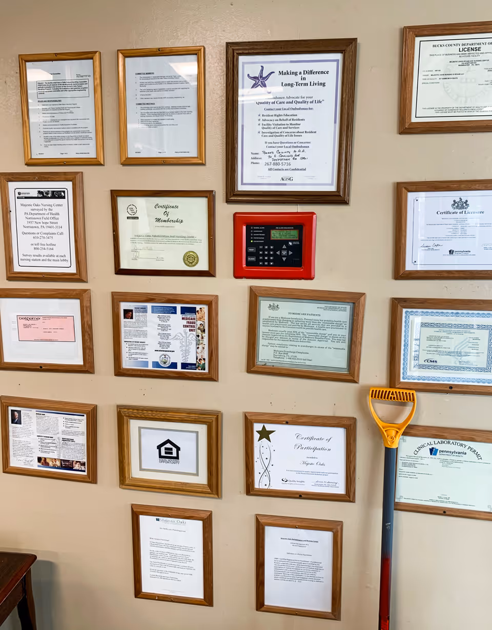 A beige wall displaying multiple framed certificates, licenses, and informational documents arranged in a grid pattern. A red fire alarm control panel is mounted in the center of the wall. A yellow and gray handled tool, possibly a snow or ice scraper, leans against the wall on the right side.