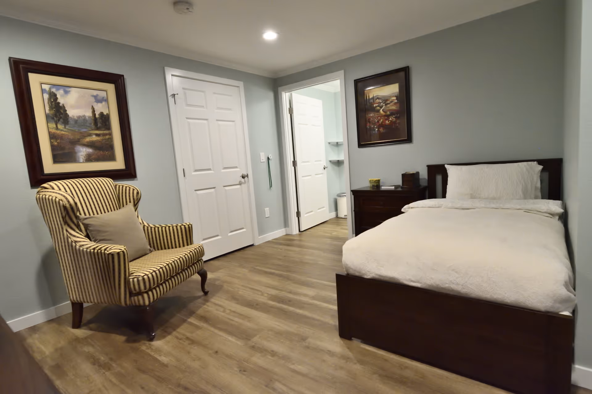 Tidy single-occupancy bedroom with a bed, striped armchair, nightstand, framed artwork, and an open doorway to a bathroom.