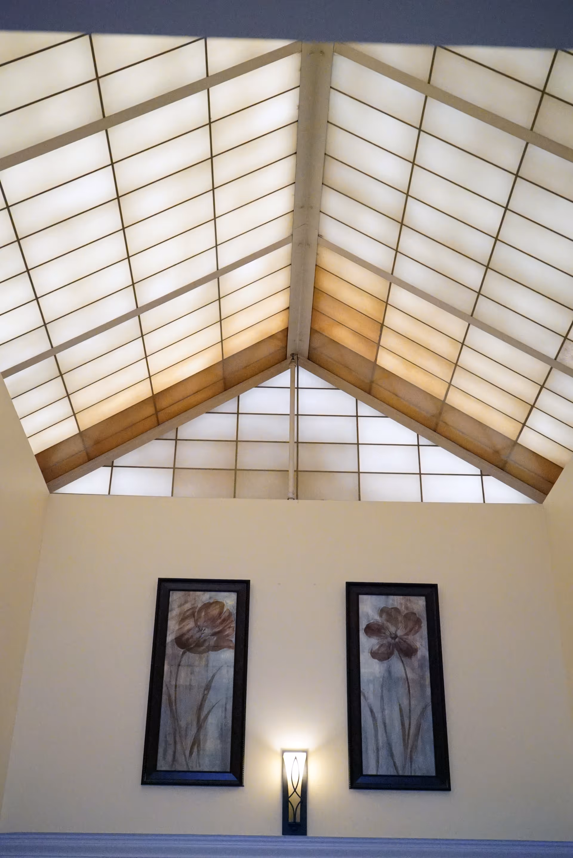 Vaulted interior with a large translucent skylight, two framed floral paintings and a lit wall sconce on the wall below.