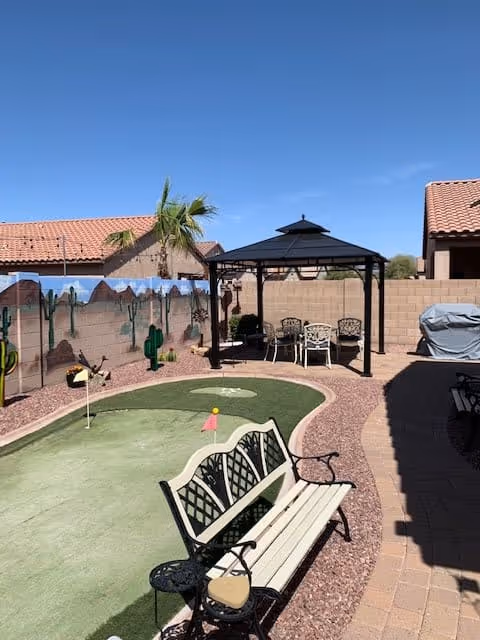 Outdoor patio area with a small putting green, a white bench with black metal armrests, a black gazebo with a table and chairs underneath, a barbecue grill covered with a gray cover, and a mural painted on a brick wall featuring desert scenery with cacti and mountains under a clear blue sky.