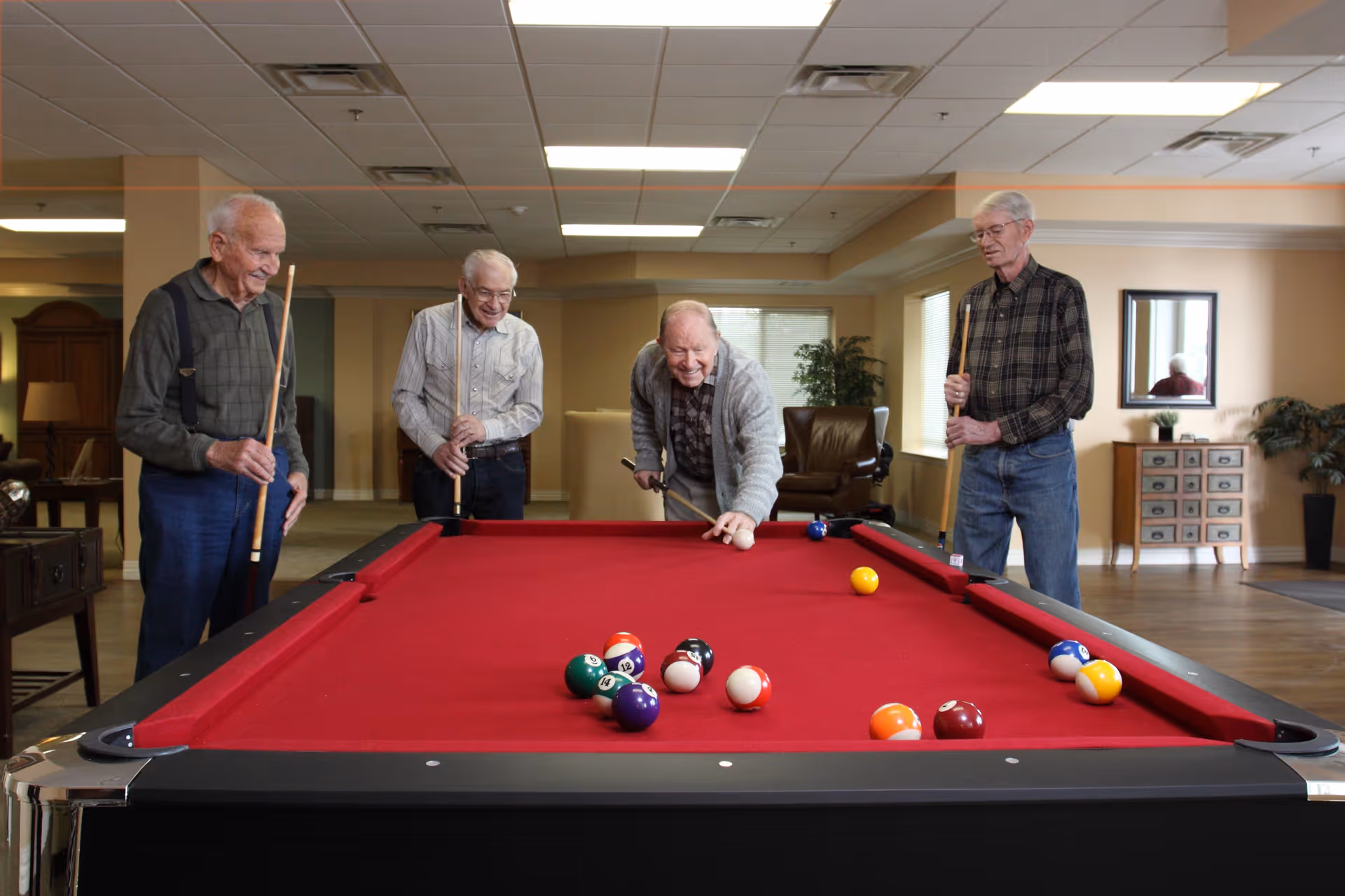 Four elderly men playing pool on a red felt pool table in a well-lit common room with wooden flooring, chairs, plants, and a mirror on the wall.