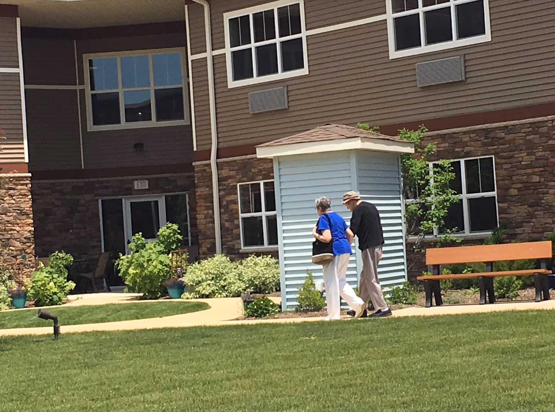 An elderly couple walking on a paved path in a well-maintained garden area outside a senior living facility building. The building has brown siding with stone accents and multiple windows. There is a wooden bench and some small shrubs and plants along the path.