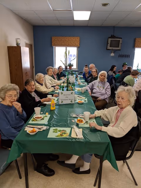 A group of elderly people sitting around a long table covered with a green tablecloth, enjoying a meal together in a dining room. The table is set with plates, cups, and condiments, and there is a sign in the center that reads 'Reserved for Woodland Acres'. The room has blue and beige walls, windows with valances, and a wall-mounted television.