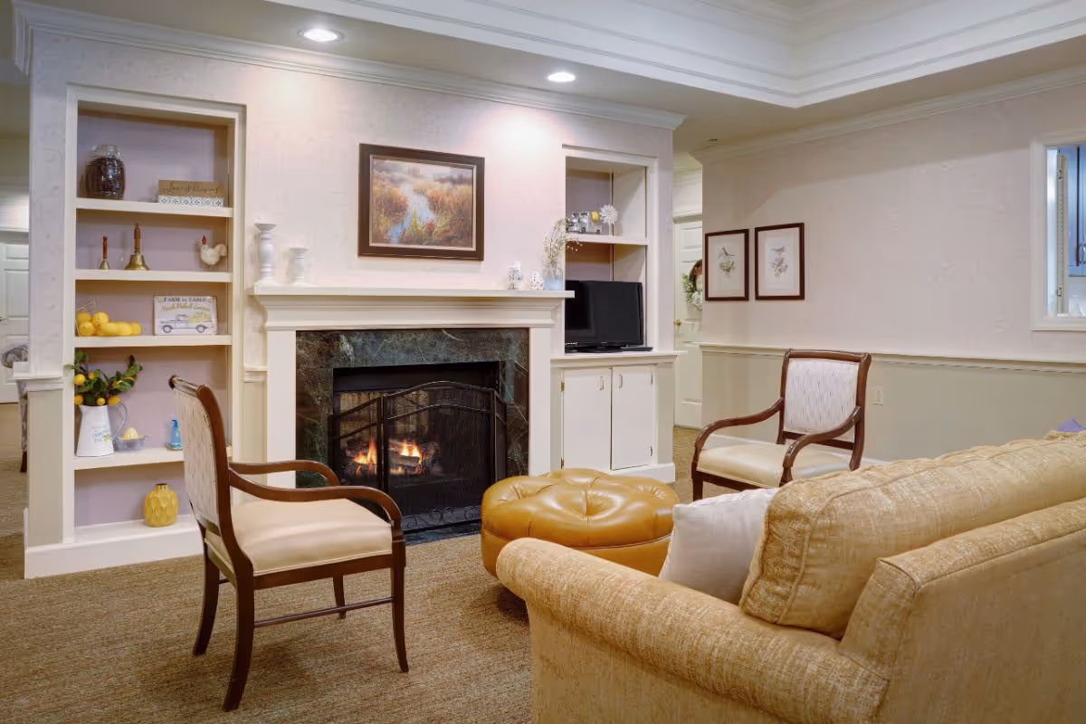 A cozy living room area in a senior living facility featuring a lit fireplace with a dark marble surround, a beige sofa with a white pillow, two wooden armchairs with cream cushions, and a round tan leather ottoman. Built-in shelves flank the fireplace, decorated with various items including a small TV, vases, and decorative signs. The walls are light-colored with framed artwork and soft lighting from recessed ceiling lights.