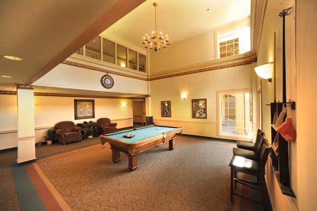 A spacious, well-lit recreational room with a pool table in the center. The room features high ceilings with a chandelier, two armchairs with a small table and plants against one wall, framed artwork on the walls, and several chairs lined up near a window. There is also a rack holding pool cues and a red hat on the right side of the image.