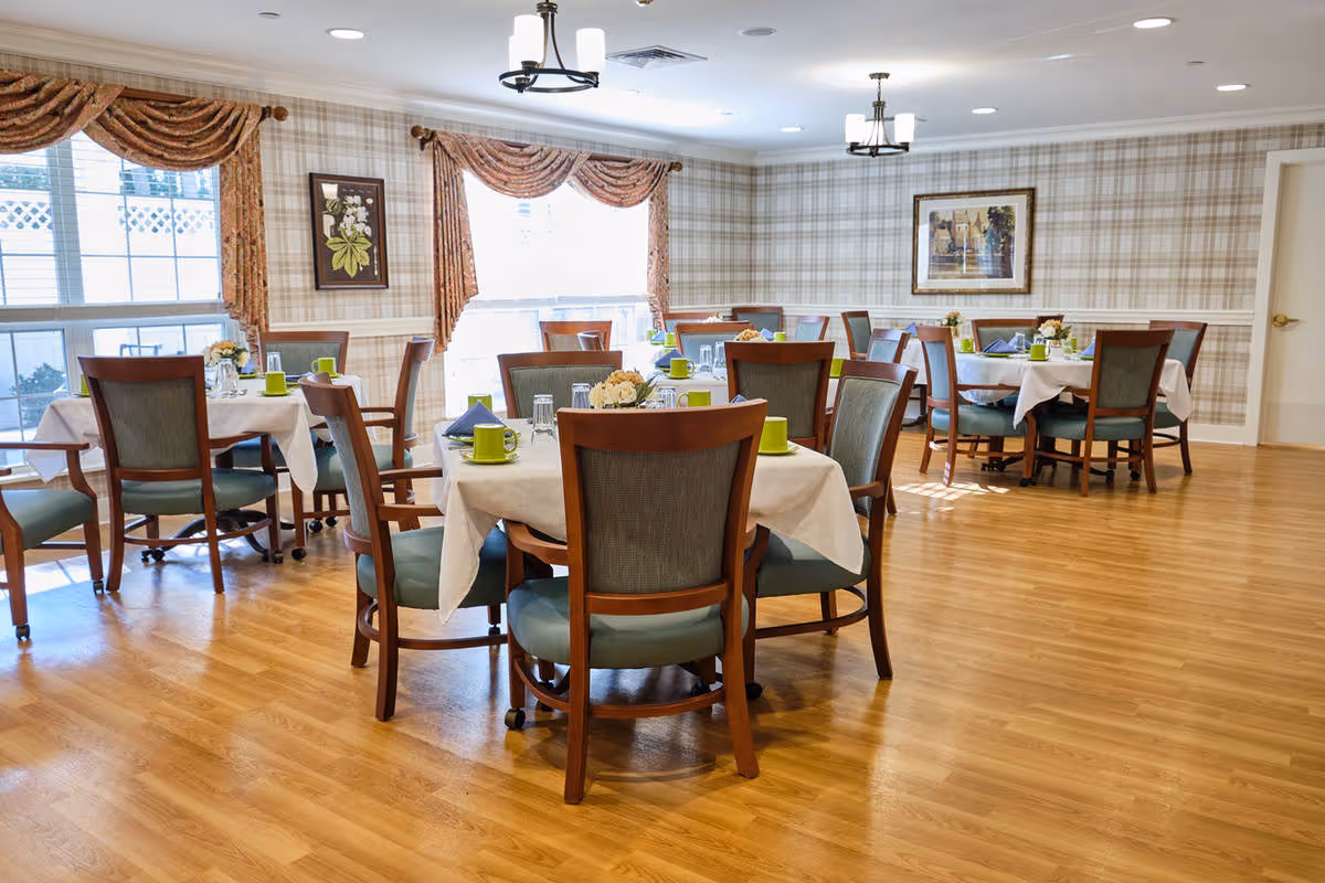 A bright dining room with several round tables covered with white tablecloths, each set with green cups, glasses, napkins, and small floral centerpieces. The room has wooden chairs with green cushions, large windows with patterned curtains, plaid wallpaper, wooden flooring, and ceiling light fixtures.