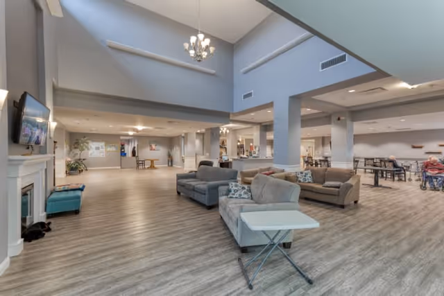 Spacious senior living facility common area with high ceilings, multiple seating arrangements including sofas and chairs, a wall-mounted TV above a fireplace, and a dining area with tables and chairs in the background. The room features light-colored walls and wood-style flooring.