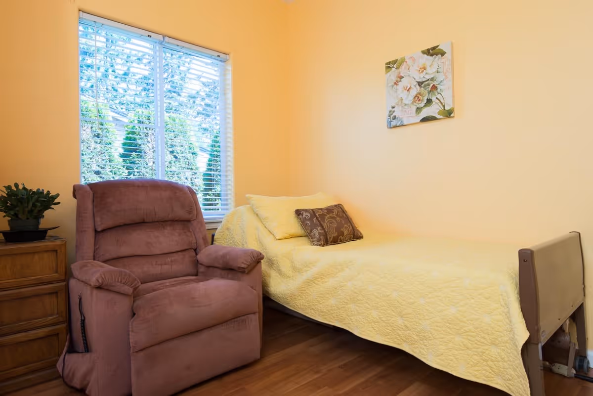 A small bedroom with a single bed covered in a yellow quilt and a brown decorative pillow. Next to the bed is a brown recliner chair and a wooden nightstand with a small potted plant. A window with white blinds lets in natural light, and a floral painting hangs on the yellow wall above the bed.