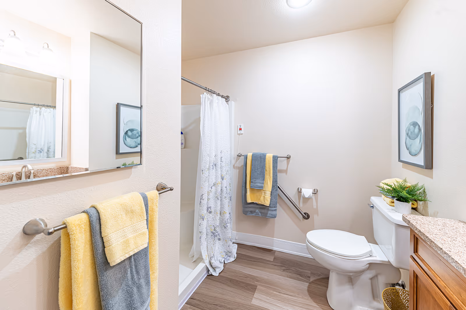 A clean and bright bathroom featuring a shower with a white floral curtain, a toilet with a small plant and yellow towels on top, a wooden vanity with a granite countertop, and two towel racks holding yellow and gray towels. The walls are light beige, and there is a framed abstract artwork hanging above the toilet.