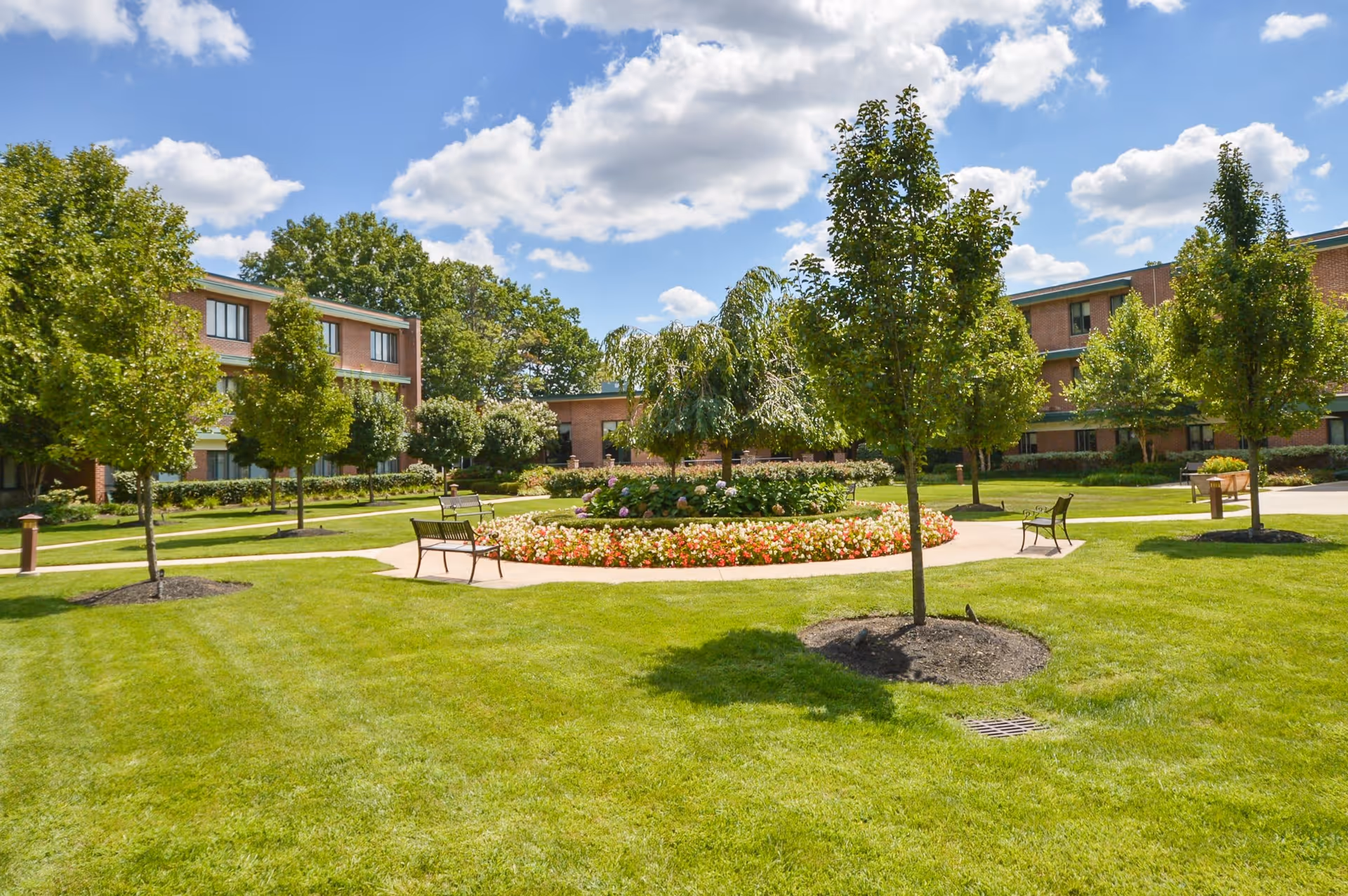 A well-maintained outdoor garden area at Wesley Enhanced Senior Living featuring green grass, several trees, a circular flower bed with colorful flowers, benches along a paved walkway, and a clear blue sky with some clouds.