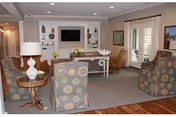 A cozy living room in an assisted living facility featuring patterned armchairs, a wooden side table with a lamp, a console table with decorative items, a wall-mounted flat screen TV, and French doors leading outside. The room has a warm and inviting atmosphere with neutral tones and soft lighting.