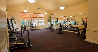 A spacious fitness room with exercise equipment including weight machines and stationary bikes. The room has large windows allowing natural light to enter, a wall-length mirror, and several ceiling lights. There are also some stacked chairs in the corner.