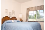 A simple bedroom with a made bed featuring a blue quilt, wooden headboard, bedside lamp, framed art on the wall, and a window with curtains.