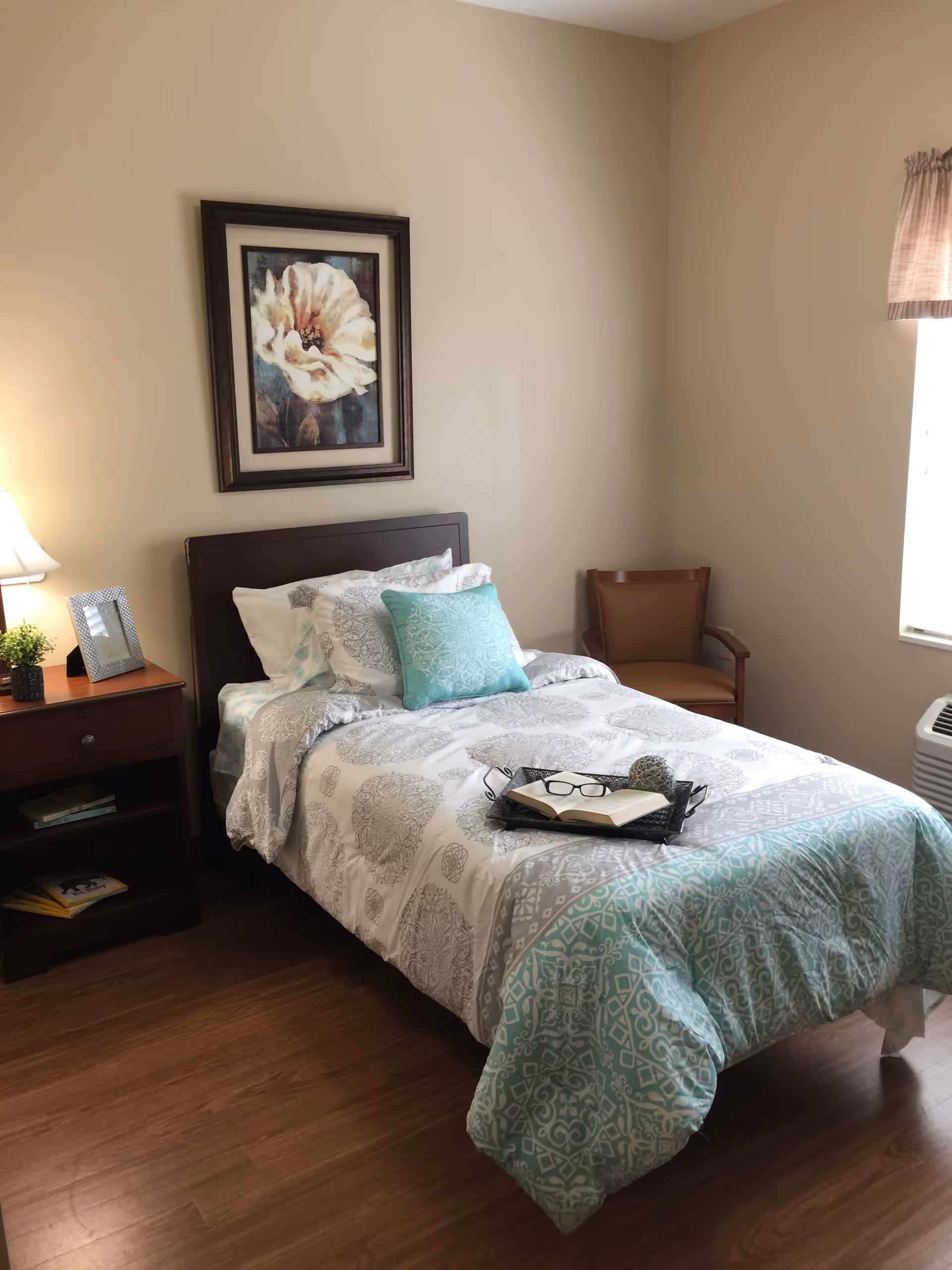A neatly made single bed with teal and gray patterned bedding beside a nightstand, chair, wall art, and a window in a bedroom.