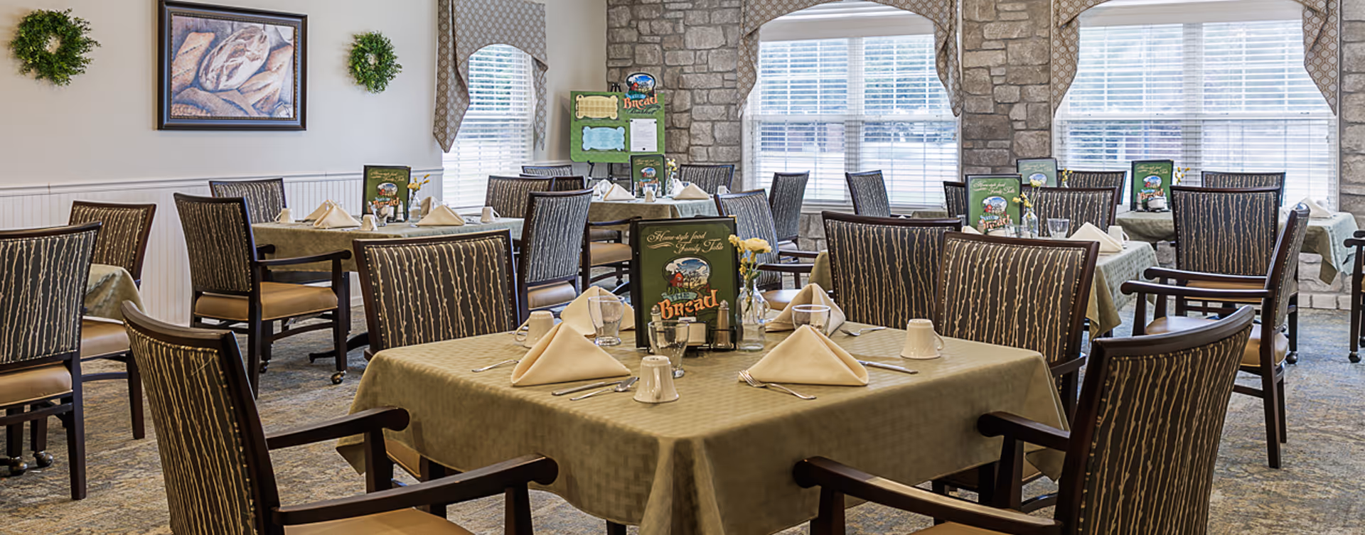 Well-lit dining room with multiple tables set with napkins, place settings and menus surrounded by upholstered chairs and large windows.