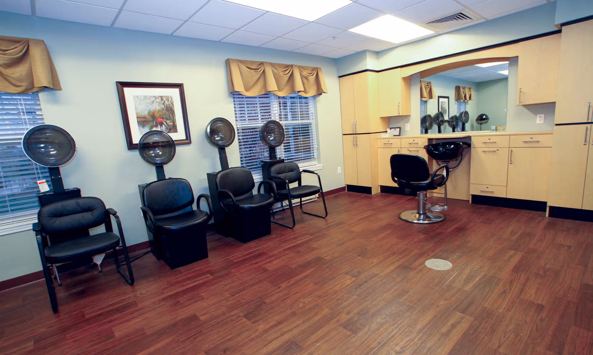 Empty salon room with hooded hair dryers, a styling chair, and cabinets along a mirrored wall.