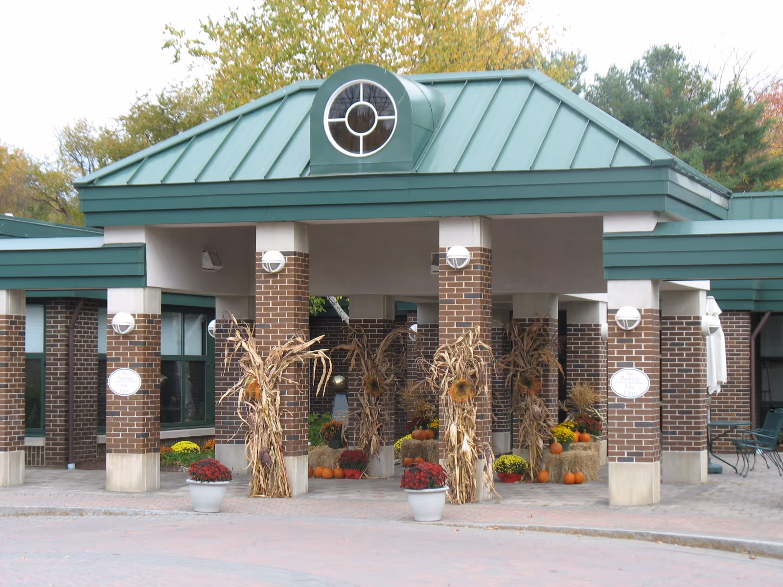 Entrance area of a building with brick pillars and a green metal roof. The entrance is decorated with dried cornstalks, pumpkins, hay bales, and colorful potted flowers, suggesting an autumn or harvest theme.