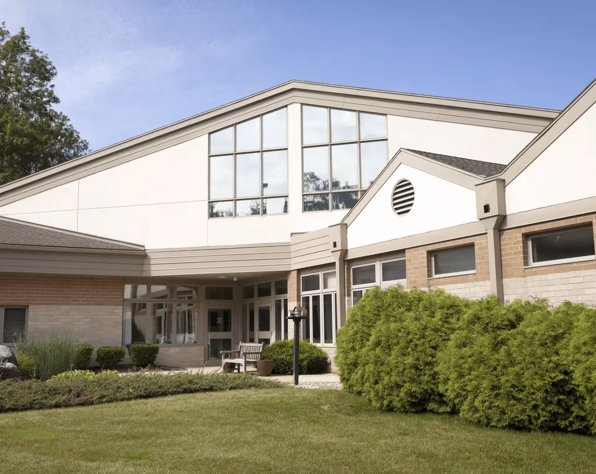 Exterior view of StoryPoint Kalamazoo (Bronson Place) building showing a well-maintained lawn, shrubs, and a bench near the entrance under a clear blue sky.
