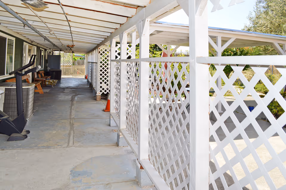 Covered outdoor walkway with white lattice fencing on the right side and a row of windows and doors on the left side. There are some chairs, an exercise machine, and other items along the left wall. The area is well-lit with natural light and has a concrete floor.